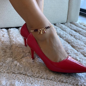 Puede incluir: Una tobillera dorada con pequeños abalorios y cuentas rojas y negras se usa con tacones altos rojos. La tobillera tiene un pequeño dije de ojo rojo y otros dijes dorados. Los zapatos son de charol.