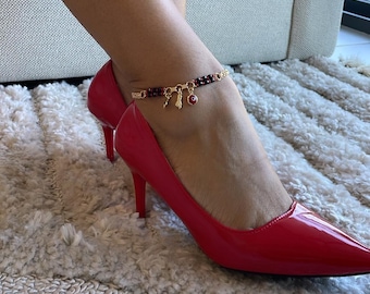Puede incluir: Una tobillera dorada con pequeños abalorios y cuentas rojas y negras se usa con tacones altos rojos. La tobillera tiene un pequeño dije de ojo rojo y otros dijes dorados. Los zapatos son de charol.