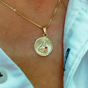 May include: A gold chain necklace with a round pendant featuring a detailed design and a small red gemstone.