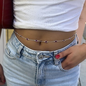 Pode incluir: Uma corrente de corpo prateada com pedras preciosas vermelhas e redondas. A corrente é usada sobre um top branco e jeans azul claro. As pedras preciosas são espaçadas uniformemente ao longo da corrente.