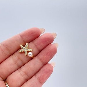 May include: A gold-colored ring featuring a starfish design adorned with clear crystals and a small white pearl. The ring is set against a light background, showcasing its delicate details and coastal theme.