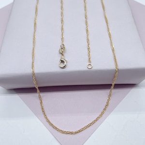 May include: A gold-colored necklace with a twisted rope chain design. The necklace features a lobster clasp closure and an adjustable chain. The jewelry is displayed on a light pink surface.