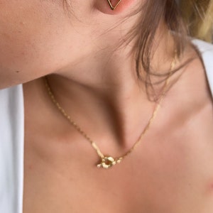 May include: A gold necklace with a knot-shaped pendant. The necklace is on a person's chest.