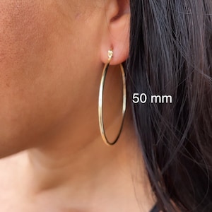 以下が含まれることがあります： 直径50mmのゴールドフープイヤリング。イヤリングは人の耳につけられています。