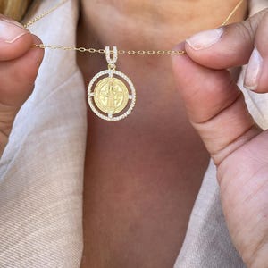 Può includere: Una collana con pendente dorato con un ciondolo circolare raffigurante una figura religiosa e cristalli. Il pendente è appeso a una delicata catena d'oro. La collana è tenuta da una persona.