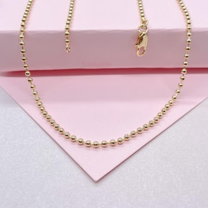 Può includere: Una collana a catena d'oro con un design a catena a palline. La catena è su una superficie rosa.