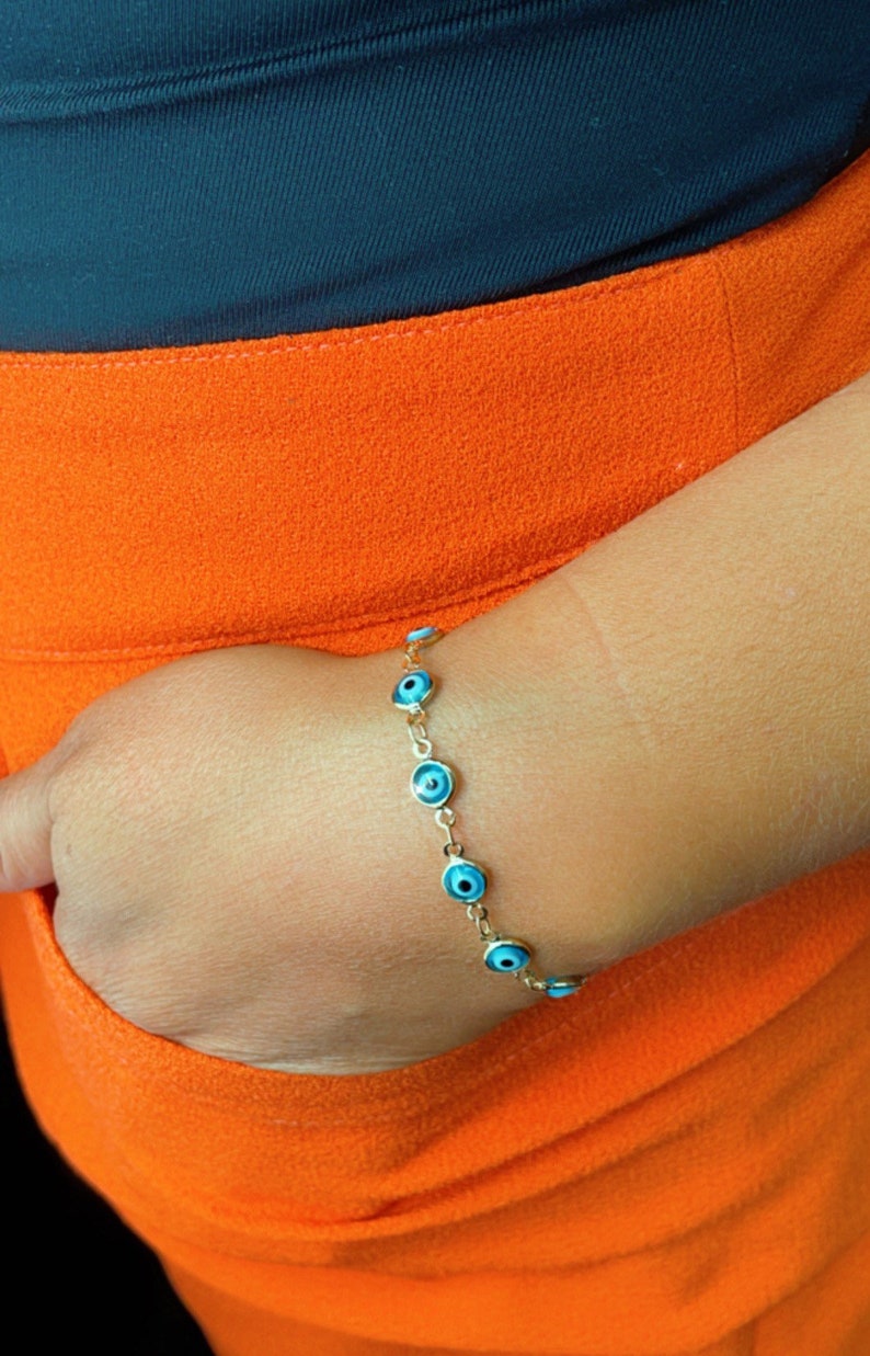 May include: A gold chain bracelet with blue evil eye charms. The bracelet is on a person's wrist.