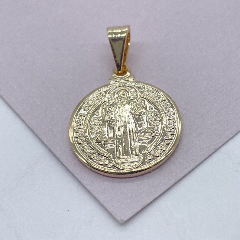 K&ouml;nnte beinhalten: Ein goldfarbener Anh&auml;nger mit einem detaillierten kreisf&ouml;rmigen Design. Der Anh&auml;nger zeigt das Bild einer religi&ouml;sen Figur und ist an einer kleinen Schlaufe aufgeh&auml;ngt. Der Anh&auml;nger ist wahrscheinlich aus goldfarbenem Metall gefertigt und wird auf einer hellvioletten Oberfl&auml;che pr&auml;sentiert.