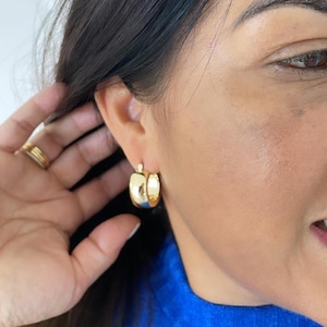 May include: Gold hoop earrings with a thick, rounded design. The earrings are worn by a person with dark hair.