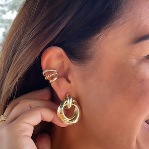 May include: Gold hoop earrings with a thick, twisted design. The earrings are worn in the earlobe and are a statement piece.