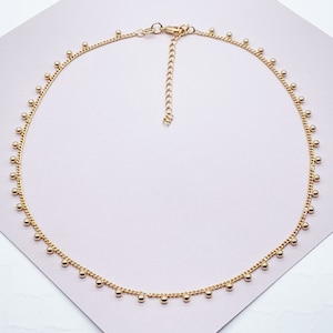 May include: A delicate gold-colored necklace featuring a fine chain adorned with small, evenly spaced spherical beads. The necklace has an adjustable length and a lobster clasp closure, suitable for various neck sizes. The jewelry is displayed on a light pink surface.
