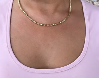 May include: A gold beaded necklace is worn around the neck. The necklace is made of small, round beads. The person is wearing a light pink scoop neck top. The person is smiling, and wearing gold stud earrings.