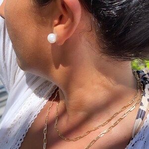 Pode incluir: Uma mulher vestindo uma blusa branca com um colar de corrente dourada. O colar &eacute; composto por duas correntes sobrepostas. A mulher tamb&eacute;m est&aacute; usando um brinco de p&eacute;rola branca.