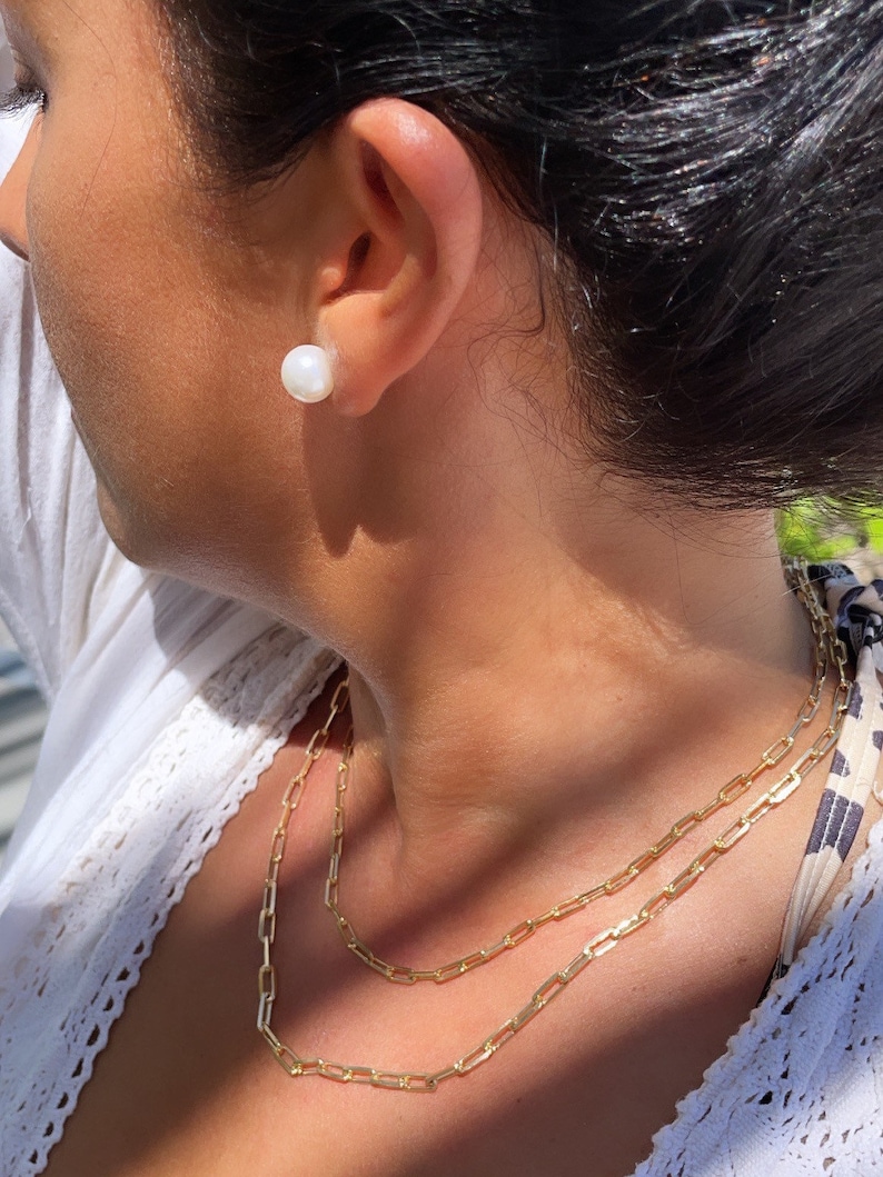 Pode incluir: Uma mulher vestindo uma blusa branca com um colar de corrente dourada. O colar &eacute; composto por duas correntes sobrepostas. A mulher tamb&eacute;m est&aacute; usando um brinco de p&eacute;rola branca.