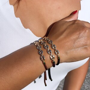 May include: Three gold chain bracelets with black, brown, and purple cords. The bracelets are stacked on a person's wrist.