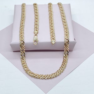 May include: Two gold chains, one longer than the other, are laid out on a pink surface. The chains are made of a thick, curb link design.