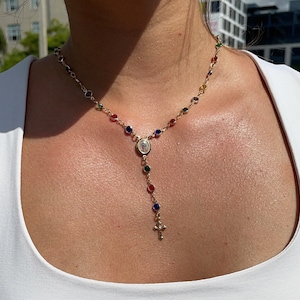 May include: Gold-coloured rosary necklace with colourful beads and a cross pendant. The necklace has a central medallion with a religious image. The beads are in various colours, including red, blue, green, and yellow. The necklace is worn around the neck.