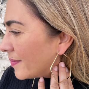 May include: Gold square hoop earrings with a slightly rounded edge. The earrings are worn by a person with blonde hair.