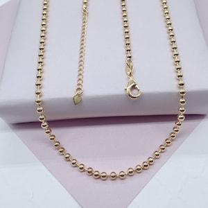 May include: A gold chain necklace with a delicate ball chain design. The chain is on a pink background.