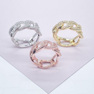 May include: Three chain-link rings in silver, gold, and rose gold. Each ring features a chain design encrusted with small, clear gemstones. The rings are displayed on a light pink surface, showcasing their different metallic finishes.