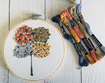 May include: A white embroidery hoop with a colorful embroidered design of four trees. The trees are in shades of orange, green, gray, and brown. The embroidery hoop is surrounded by six spools of embroidery floss in various colors.