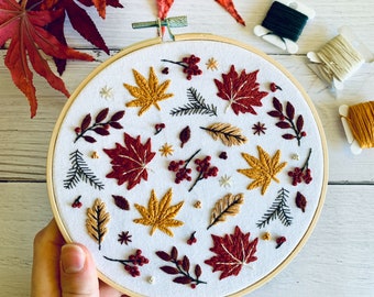 May include: A white embroidery hoop featuring a colourful autumn leaf design. The design includes red, orange, yellow and green leaves, as well as brown branches and berries. The leaves are embroidered using a variety of stitches, including satin stitch, French knots and back stitch.