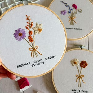 May include: Three circular wooden embroidery hoops featuring white fabric with hand-stitched floral bouquets. The primary hoop displays a purple flower, orange sprigs, and a white daffodil with the text MUMMY ELSIE DADDY EST. 2026 stitched in black.