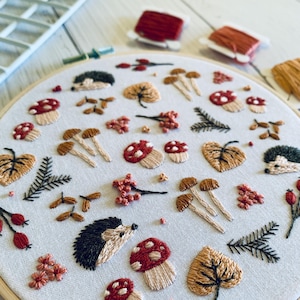 May include: An embroidered hoop art piece featuring a woodland theme. The design includes mushrooms, leaves, berries, and hedgehogs in shades of red, brown, and green. The embroidery is set against a white fabric background.