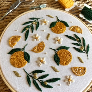 Puede incluir: Un bastidor de bordado circular de madera con tela blanca que presenta diseños cosidos a mano de naranjas amarillas, rodajas de naranja, flores blancas y hojas de color verde oscuro, realizados con hilos detallados en tonos amarillos, blancos y verdes.