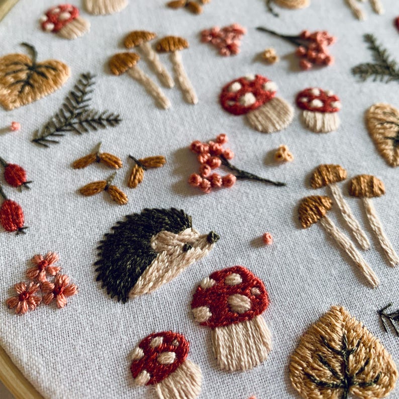 May include: Embroidered artwork featuring a woodland theme. The design includes a hedgehog, mushrooms with red and white caps, and various leaves and berries in shades of brown, beige, and red. The embroidery is on a light-colored fabric.