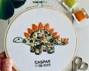 May include: Embroidered hoop art featuring a floral patterned dinosaur with orange spikes. The hoop is wooden and the fabric is white with the name "CASPAR" and the date "17.09.2022" embroidered below the dinosaur.