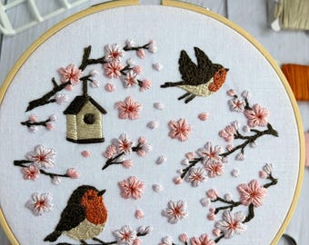 May include: Embroidered hoop art featuring a spring scene with two birds, a birdhouse, and cherry blossoms. The birds have brown and orange coloring. The blossoms are pink and white, and the branches are brown. The embroidery is set in a wooden hoop.