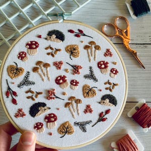 May include: An embroidery hoop featuring a white fabric with an autumn-themed design. The design includes embroidered mushrooms, leaves, berries, hedgehogs, and sprigs in shades of brown, red, and green. A pair of gold scissors and spools of thread are visible.