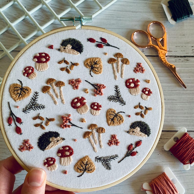 May include: An embroidery hoop featuring a white fabric with an autumn-themed design. The design includes embroidered mushrooms, leaves, berries, hedgehogs, and sprigs in shades of brown, red, and green. A pair of gold scissors and spools of thread are visible.