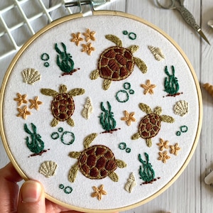 May include: Embroidered hoop art with sea turtles, starfish, and seashells. The turtles have brown and gold shells, with green seaweed and bubbles. The design is on a white background, framed by a wooden hoop.
