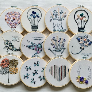 Puede incluir: Diez aros de bordado con varios diseños, que incluyen un globo aerostático, una bombilla, un par de botas, un colibrí, una regadera, una raya, un árbol, un corazón, una herradura y un grupo de flores. Los aros son todos de madera y tienen un fondo blanco.