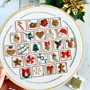 May include: A hand-stitched Christmas advent calendar in a white hoop. The calendar has 24 numbered squares with festive designs, including a snowman, Christmas tree, and candy cane. The embroidery uses red, green, and gold thread.