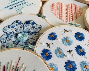 May include: A collection of embroidery hoops with various designs, including a blue star, a pink heart, a blue flower, a blue clover, and a blue floral pattern with bees. The hoops are made of wood and have a white fabric background.