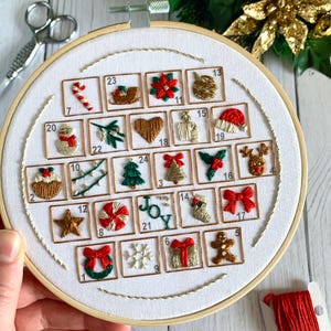 May include: Embroidered Christmas advent calendar in a white hoop. Each square features a festive design, including a candy cane, Christmas tree, and reindeer. The word "JOY" is embroidered in green. The hoop measures approximately 8 inches in diameter.