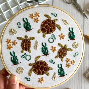 May include: Embroidered hoop art featuring a whimsical underwater scene with multiple brown and green sea turtles, seaweed, seashells, and starfish. The hoop is made of wood and has a white fabric background.