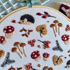 May include: Embroidered artwork featuring a woodland theme. The design includes a hedgehog, mushrooms, leaves, and berries, all in shades of brown, red, and green. The embroidery is set within a wooden hoop, creating a framed piece.