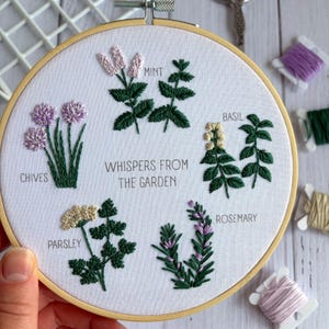 Puede incluir: Arte bordado en bastidor con hierbas: cebollino, menta, albahaca, perejil y romero. El texto "Whispers From The Garden" está centrado. El bordado utiliza hilo verde, morado y crema sobre tela blanca, en un bastidor de madera.