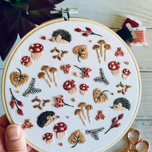 Peut inclure: Un cerceau de broderie blanc avec un motif d'automne coloré. Le motif représente des hérissons, des champignons, des feuilles et des baies dans des tons de rouge, marron, or et vert.