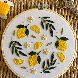 Peut inclure: Broderie sur tambour représentant des citrons jaunes, des tranches de citron, des fleurs blanches et des feuilles vertes. Le motif est cousu sur du tissu blanc et monté dans un cercle en bois. Une bobine de fil jaune et des ciseaux sont en arrière-plan.