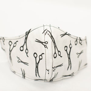 May include: White face mask with a repeating pattern of black scissors. The mask is made of fabric and has a curved shape to fit the face. The scissors are in various sizes and orientations.