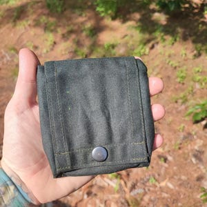 Puede incluir: Una bolsa cuadrada de color verde oscuro con un cierre de botón único, redondo y plateado. La bolsa está hecha de una tela texturizada y presenta costuras visibles en los bordes. El fondo es una escena borrosa al aire libre con follaje verde.