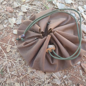 Puede incluir: Una bolsa de cordón marrón con un cordón verde y un cierre de madera. La bolsa está recogida en la parte superior y tiene una forma redonda.