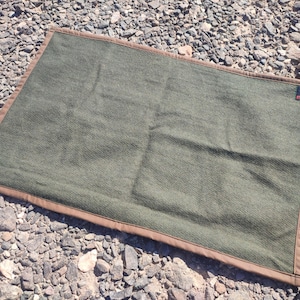 Puede incluir: Una manta de lana verde con ribete de cuero marrón. La manta tiene una pequeña etiqueta verde con la palabra "Green" en ella.