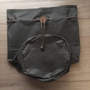 Könnte beinhalten: Eine dunkelgrüne Canvas-Zugbeutel mit einem Ledergriff und einem runden Boden. Die Tasche ist geschlossen und liegt auf einer Holzoberfläche.