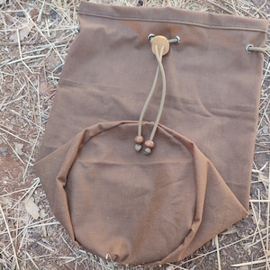 Könnte beinhalten: Eine braune Canvas-Zugschnursack mit einem Holz-Knebel und braunen Lederakzenten. Die Tasche ist gefaltet und liegt auf einem Bett aus trockenem Gras.